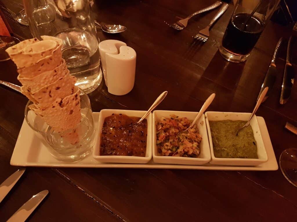 A rectangular platter holds three small bowls containing chutney, a vegetable mix, and green sauce, each with a spoon. On the left, rolled poppadom crisps are stacked upright in a glass. Cutlery, glasses, a jug, and a salt shaker are on the dark wooden table.