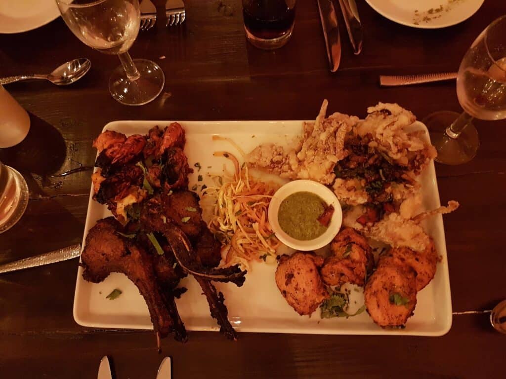A rectangular white plate with grilled lamb chops, tandoori chicken, prawns, fried calamari, shredded salad, and a small bowl of green chutney. Wine glasses, cutlery, and drinks surround the plate on a dark wooden table.