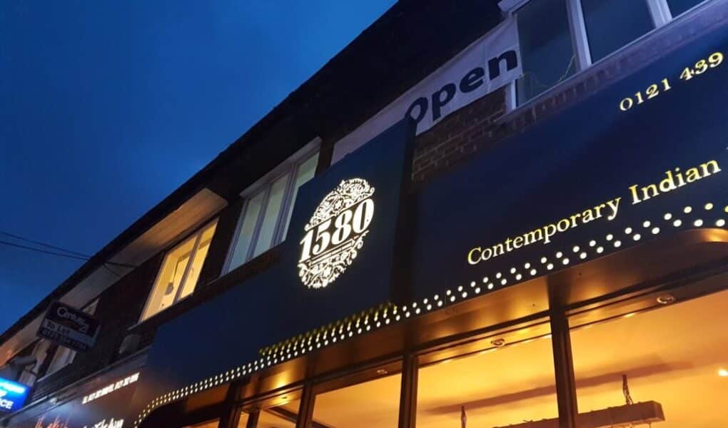 A blue evening sky above the exterior of a restaurant called "1580 Contemporary Indian." The illuminated sign features ornate detailing around “1580.” Large windows and glowing lights can be seen inside, and a sign above reads “Open”.
