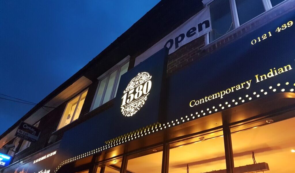 A blue evening sky above the exterior of a restaurant called "1580 Contemporary Indian." The illuminated sign features ornate detailing around “1580.” Large windows and glowing lights can be seen inside, and a sign above reads “Open”.