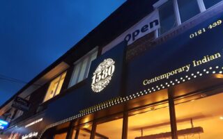 A blue evening sky above the exterior of a restaurant called "1580 Contemporary Indian." The illuminated sign features ornate detailing around “1580.” Large windows and glowing lights can be seen inside, and a sign above reads “Open”.