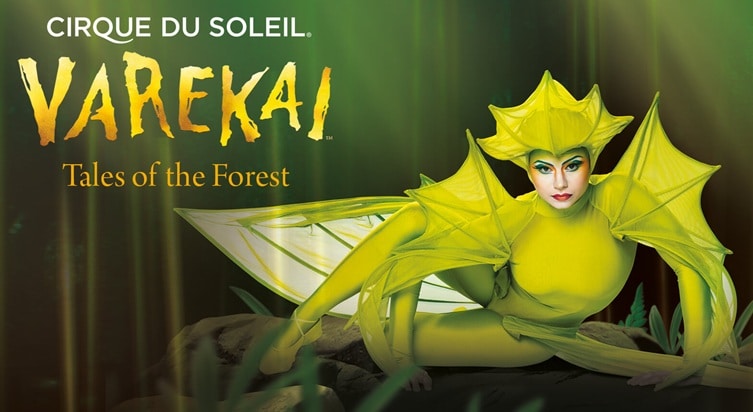 A performer in a bright yellow, leaf-inspired costume poses on a mossy surface. Text reads “CIRQUE DU SOLEIL VAREKAI Tales of the Forest.” The background is green and blurred, evoking a mystical forest setting. The performer’s make-up is dramatic and colourful.