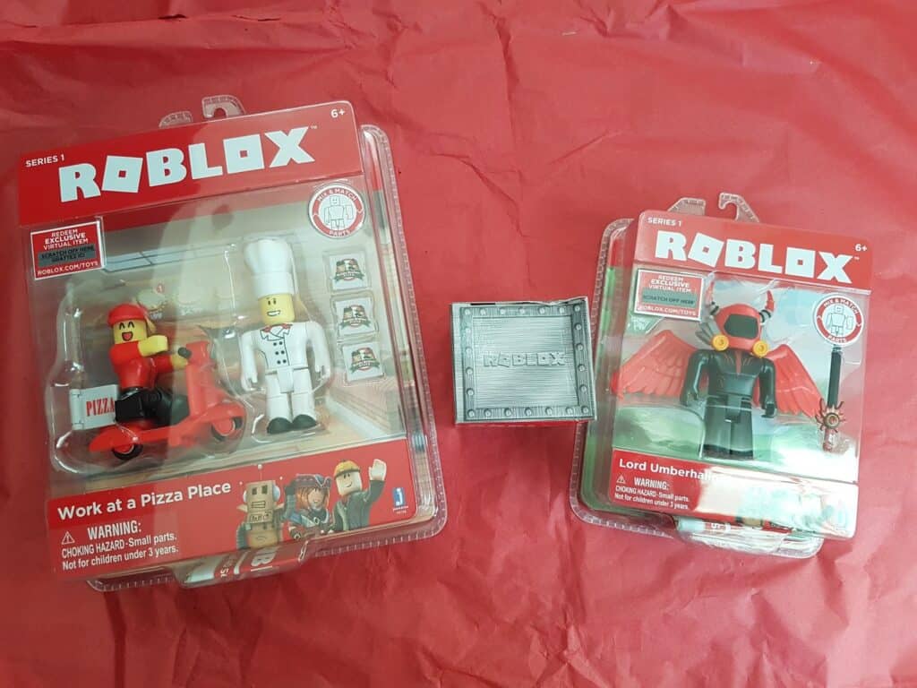 Two packaged Roblox toy sets are displayed on red paper. The left pack shows “Work at a Pizza Place” with chef and delivery figures. The right has “Lord Umberhallow” in armour with a sword. A silver Roblox collector’s box is placed between them.