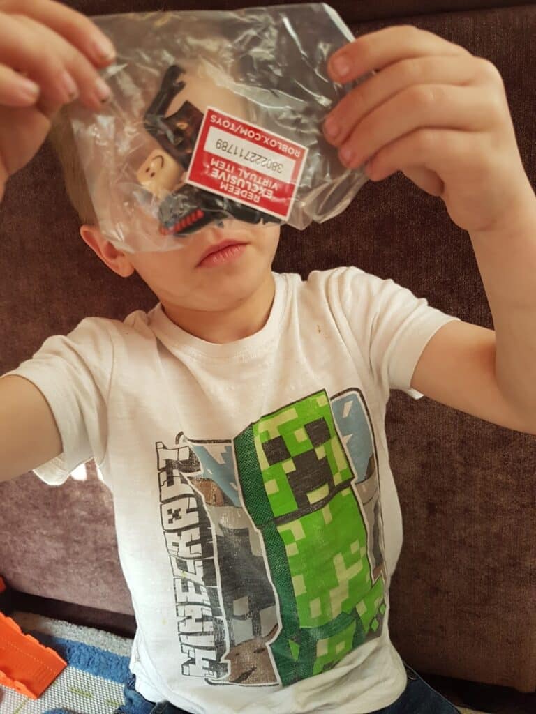A child in a white Minecraft Creeper T-shirt holds up a clear plastic bag containing LEGO minifigure pieces and a red label. The child is sitting on a brown sofa, partially obscuring their face with the bag.