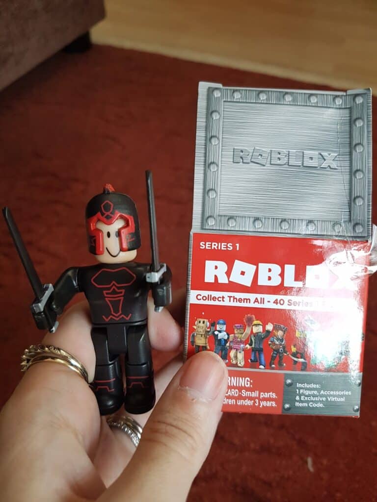 A hand holds a Roblox action figure dressed in black and red armour with two swords, alongside its grey crate-shaped box. The box shows "ROBLOX" branding, a warning label, and small images of other collectable figures from Series 1.