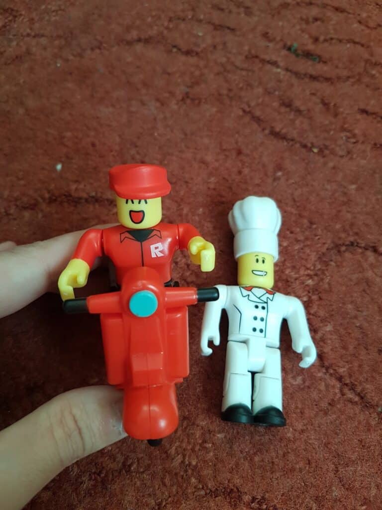 Two Roblox toy figures on a reddish carpet: one wears a red uniform and hat, riding a red scooter; the other is dressed as a chef in a white uniform and tall chef’s hat. A hand holds the scooter upright.