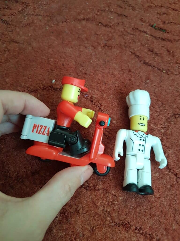 A hand holds a toy pizza delivery figure on a red scooter labelled "PIZZA" beside a white-clad chef figure, both on a textured reddish-brown surface.