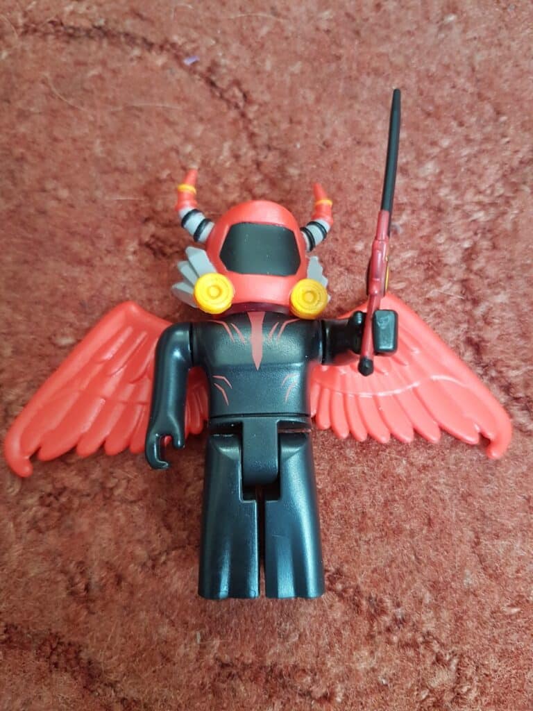 A toy figure with red wings, a black outfit, and a helmet with red and black horn-like decorations. The figure has yellow circular details on the helmet and is holding a black and red sword, lying on a brown textured surface.