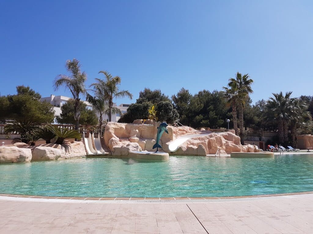 A large outdoor swimming pool with turquoise water, surrounded by palm trees and tropical plants. There are rock formations with three water slides and a dolphin statue in the centre. White sun loungers line the poolside under a clear blue sky.