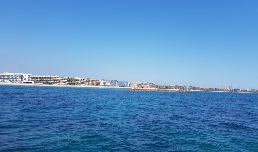 Bright blue ocean water in the foreground with gentle waves, under a clear sky. In the background, there are modern white and beige buildings lining the distant shoreline, and a small rocky breakwater extends from the coastline into the water.