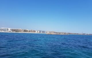 Bright blue ocean water in the foreground with gentle waves, under a clear sky. In the background, there are modern white and beige buildings lining the distant shoreline, and a small rocky breakwater extends from the coastline into the water.