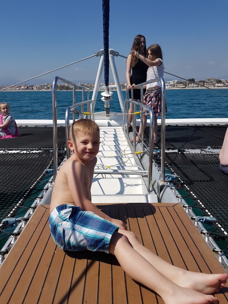 A young boy in swimming trunks lies on a wooden deck of a boat under a clear blue sky. Behind him, two girls stand together by the railings, and another girl sits alone near the edge. The sea and a distant shore are visible in the background.