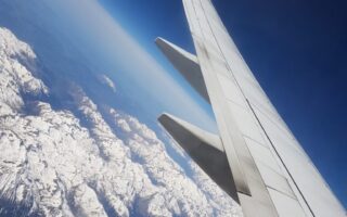 A photo taken from an aeroplane window shows a Ryanair wing in the foreground with snow-covered mountains and rugged terrain visible below, under a clear blue sky. The image captures the contrast between the aircraft wing and the dramatic landscape.