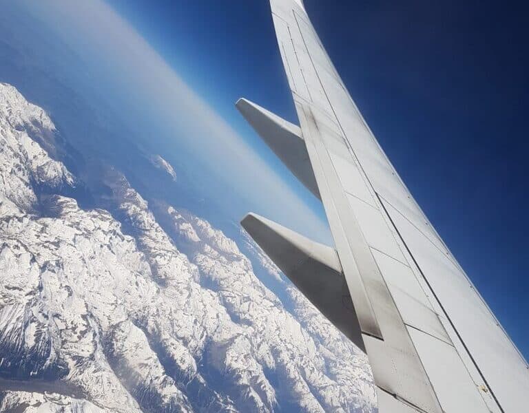 A photo taken from an aeroplane window shows a Ryanair wing in the foreground with snow-covered mountains and rugged terrain visible below, under a clear blue sky. The image captures the contrast between the aircraft wing and the dramatic landscape.