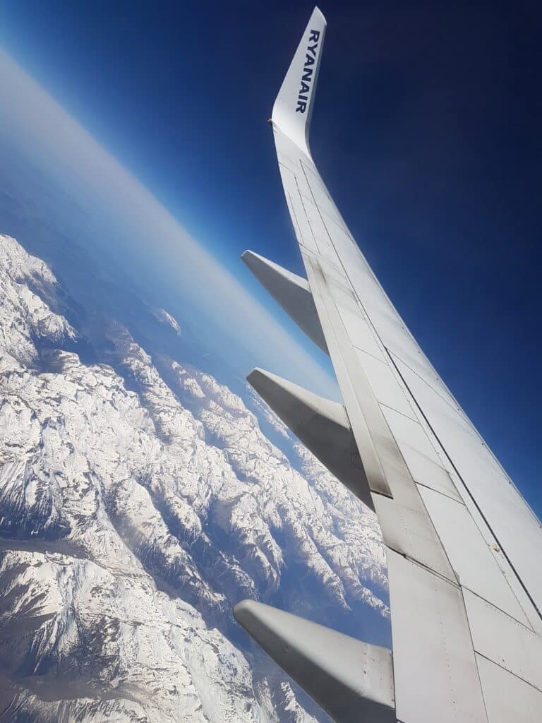 A Ryanair aeroplane wing seen from a passenger window, soaring above snow-covered mountains under a deep blue sky. The landscape below features rugged peaks and ridges, contrasting with the smooth, metallic surface of the wing.