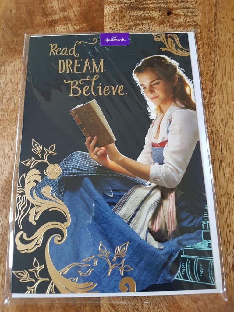 A greeting card featuring a young woman in period clothing reading a book. Gold floral designs and books decorate the border. Text reads: “Read. DREAM. Believe.” The Hallmark logo appears at the top. The background is dark, making the woman stand out.