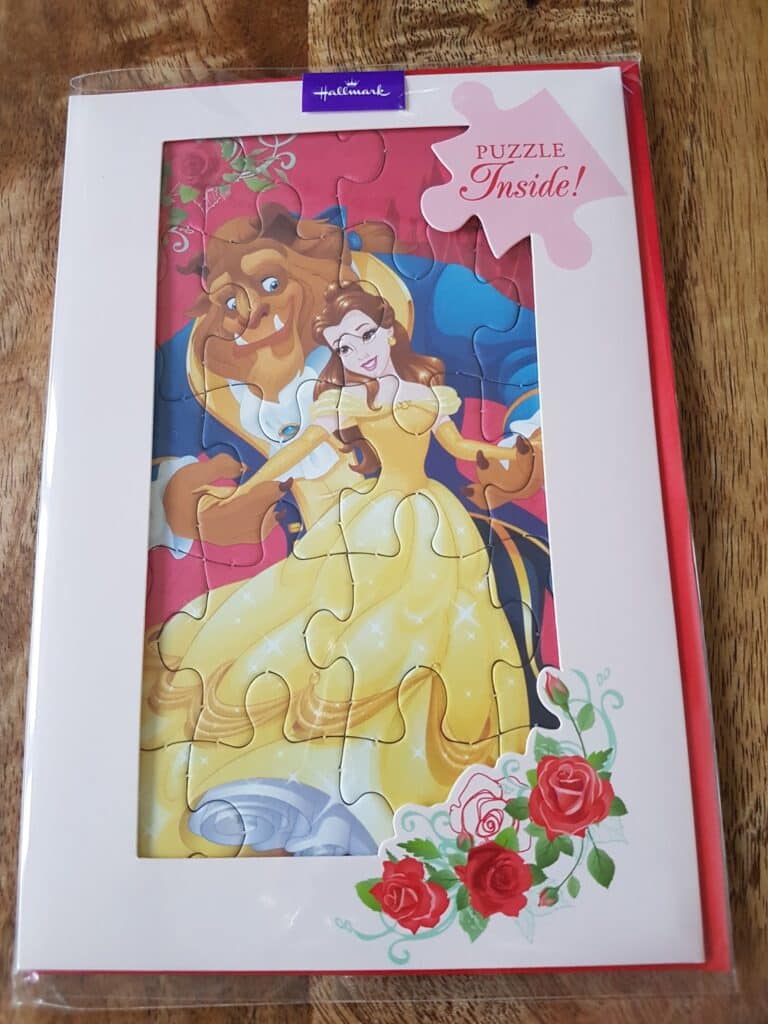 A Hallmark card features a puzzle with Disney’s Belle in a yellow dress and the Beast dancing. The card has a white border with roses at the bottom right and a pink label that says “PUZZLE Inside!” on the top right. The surface is on a wooden table.