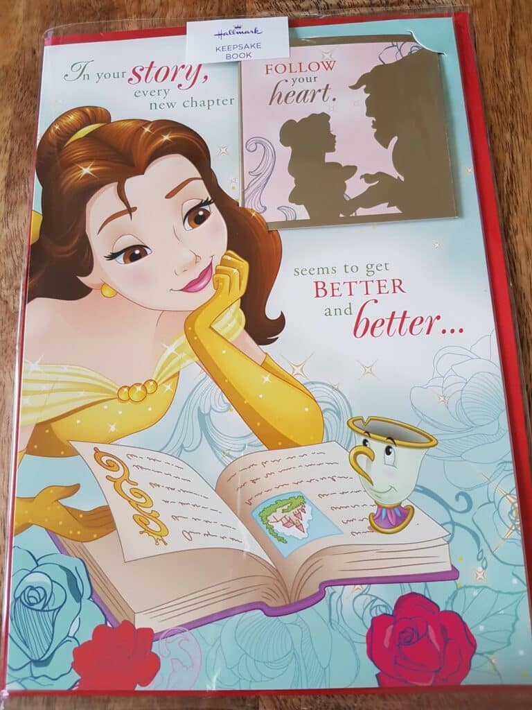 A colourful card features Belle from Disney’s "Beauty and the Beast" reading an illustrated book. Beside her is Chip, the teacup. Text reads: “In your story, every new chapter seems to get better and better…” A small card with a couple’s silhouette says: “Follow your heart.”.