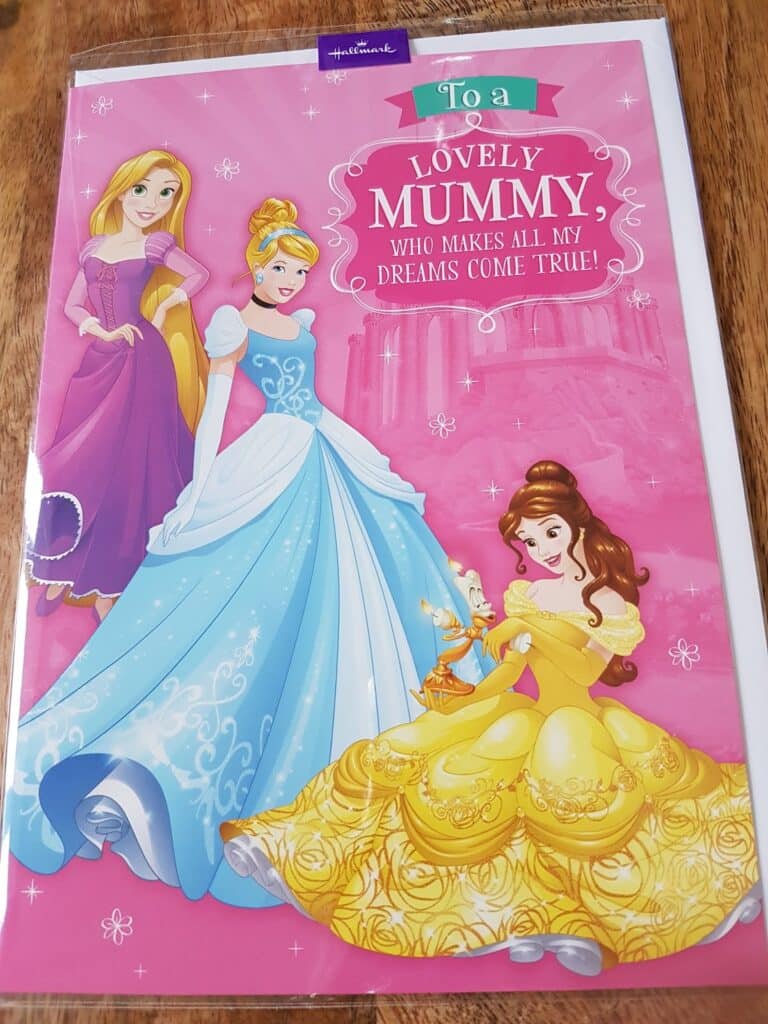 A greeting card with Disney princesses Rapunzel, Cinderella, and Belle. The background is pink with sparkles and a castle. Text reads: "To a lovely Mummy, who makes all my dreams come true!" The card is on a wooden surface.