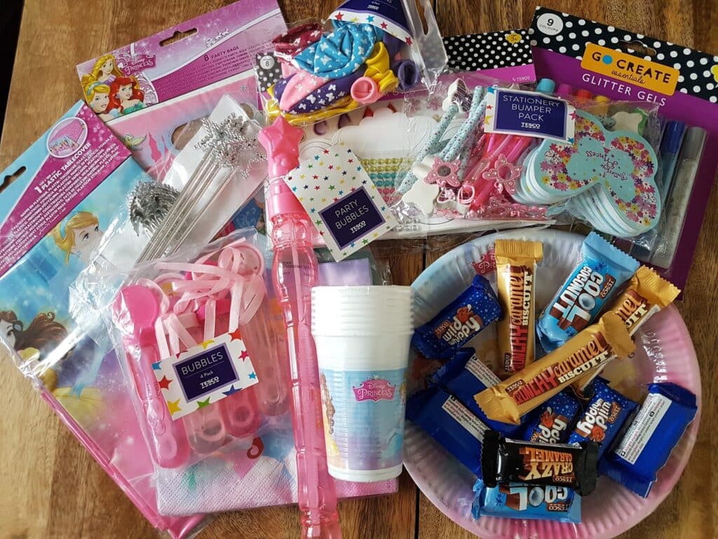 A wooden table holds assorted kids’ party supplies: packs of wands, serviettes, cups, stationery, bubble tubes, glitter gel pens, cupcake cases with blue and pink hearts, and a paper plate piled with various mini chocolate bars and sweets.