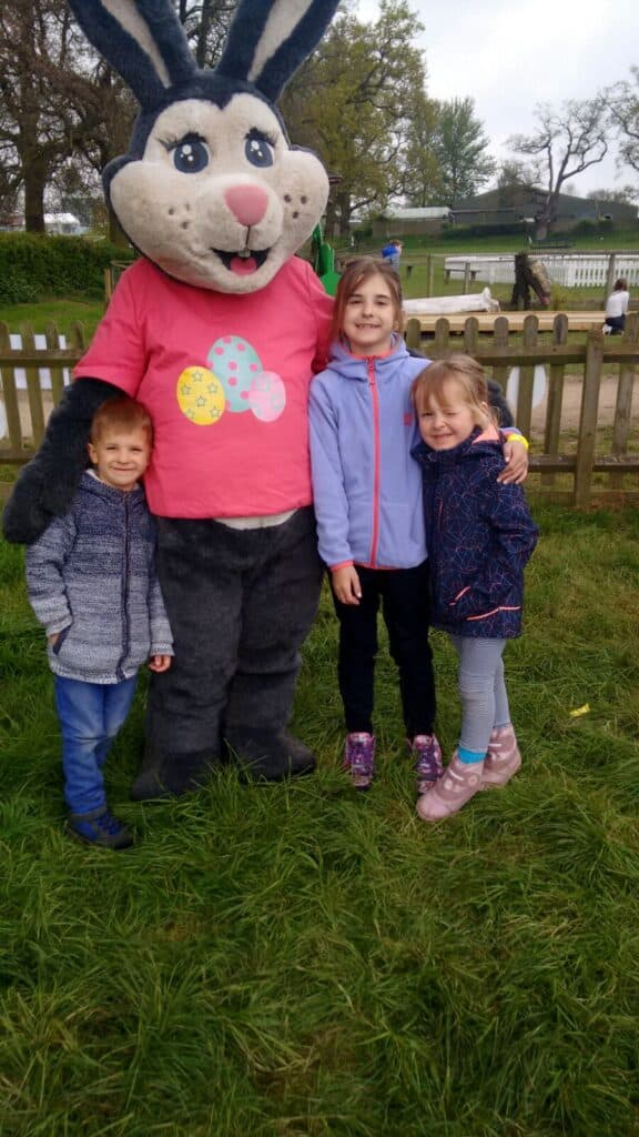 Three children stand on grass, smiling and posing with a person in a large Easter Bunny costume wearing a pink shirt with decorated eggs. A wooden fence, pond, and trees are visible in the background on a cloudy day at an outdoor event.