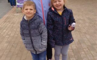 Three young children stand smiling on a paved path outside a zoo entrance. Behind them, a purple sign reads “Welcome” at the entrance to Hatters Farm Zoo. The children wear jackets and wellies, and wooden buildings can be seen in the background.