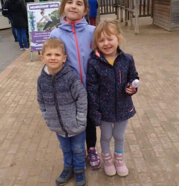 Three young children stand smiling on a paved path outside a zoo entrance. Behind them, a purple sign reads “Welcome” at the entrance to Hatters Farm Zoo. The children wear jackets and wellies, and wooden buildings can be seen in the background.