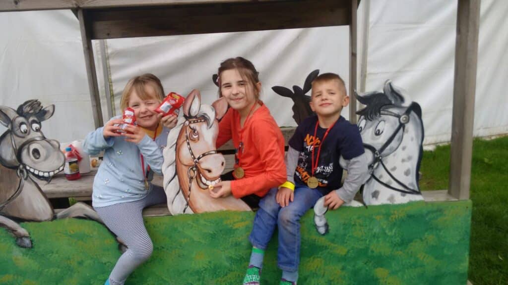 Three smiling children with medals sit and pose on a wooden structure decorated with painted horses. Two girls hold juice drinks, one boy sits beside them, all in casual clothes. The background includes a white wall and green grass.