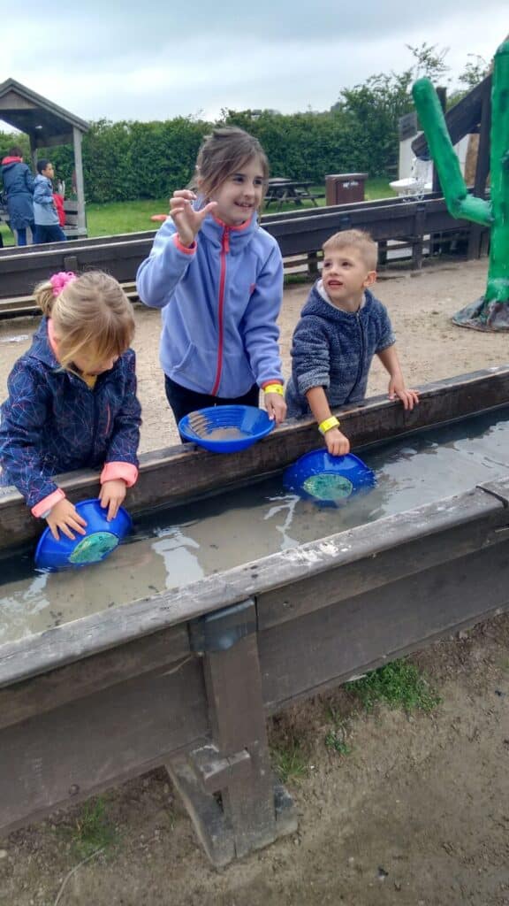 Three children play at a wooden water trough, panning for stones or gems with blue sieves. One girl in a blue jacket holds up a stone and smiles, another girl looks down at her sieve, and a young boy watches her. Other children and adults are in the background.