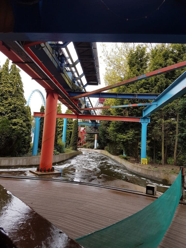 A view from a water ride at an amusement park shows an empty flume lined with green trees, red and blue metal supports above, and water pooled along the concrete channel. A rollercoaster track and loop are visible in the background through the trees.