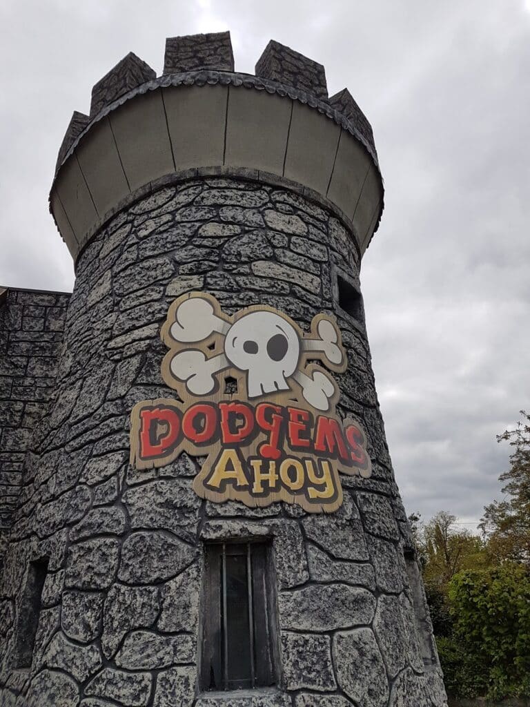 A grey, stone-textured castle tower with battlements displays a sign featuring a white cartoon skull and crossbones above the words "DODGEMS AHOY" in bold, red and yellow letters. The sky is overcast and some trees are visible in the background.