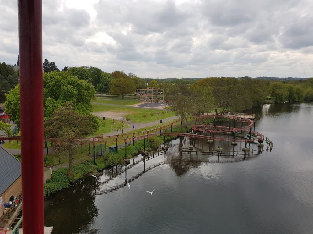 A curved red bridge crosses over a calm lake surrounded by trees and grassy areas. People walk on the paths below; a few birds are near the water. The sky is cloudy, and a partially constructed building is visible in the distance.