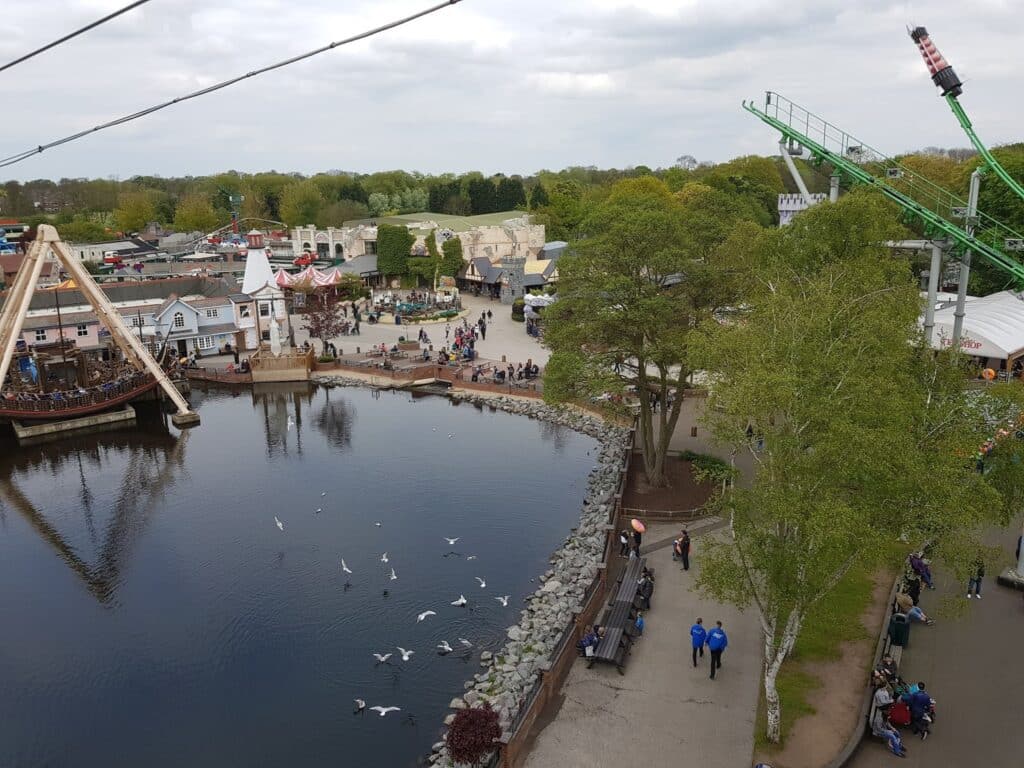 Aerial view of an amusement park with a lake, pirate ship ride on the left, scattered visitors, trees, and various buildings. Seagulls fly over the water. Rides and attractions are visible, with cloudy skies overhead. Paths wind through the park.