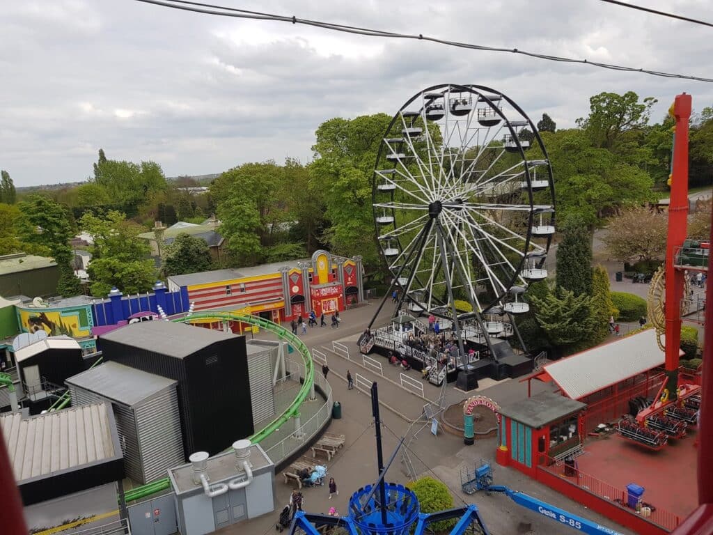 Aerial view of an amusement park featuring a large black-and-white Ferris wheel, colourful buildings, ticket booths, and green trees. People walk around the area, and the sky is overcast. Rides and attractions are scattered throughout the scene.