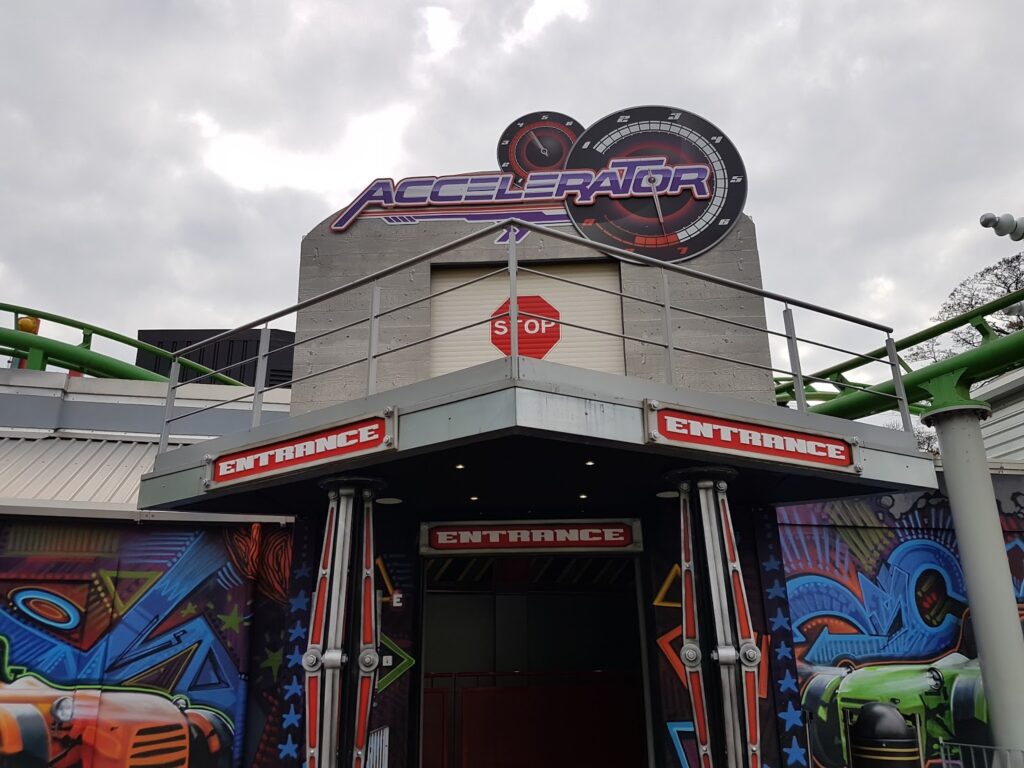 The image shows the entrance to a ride called "Accelerator" with a large futuristic sign above. The entrance is flanked by two red “ENTRANCE” signs and features bold, colourful designs and gears, with a stop sign visible above the doorway. The sky is overcast.