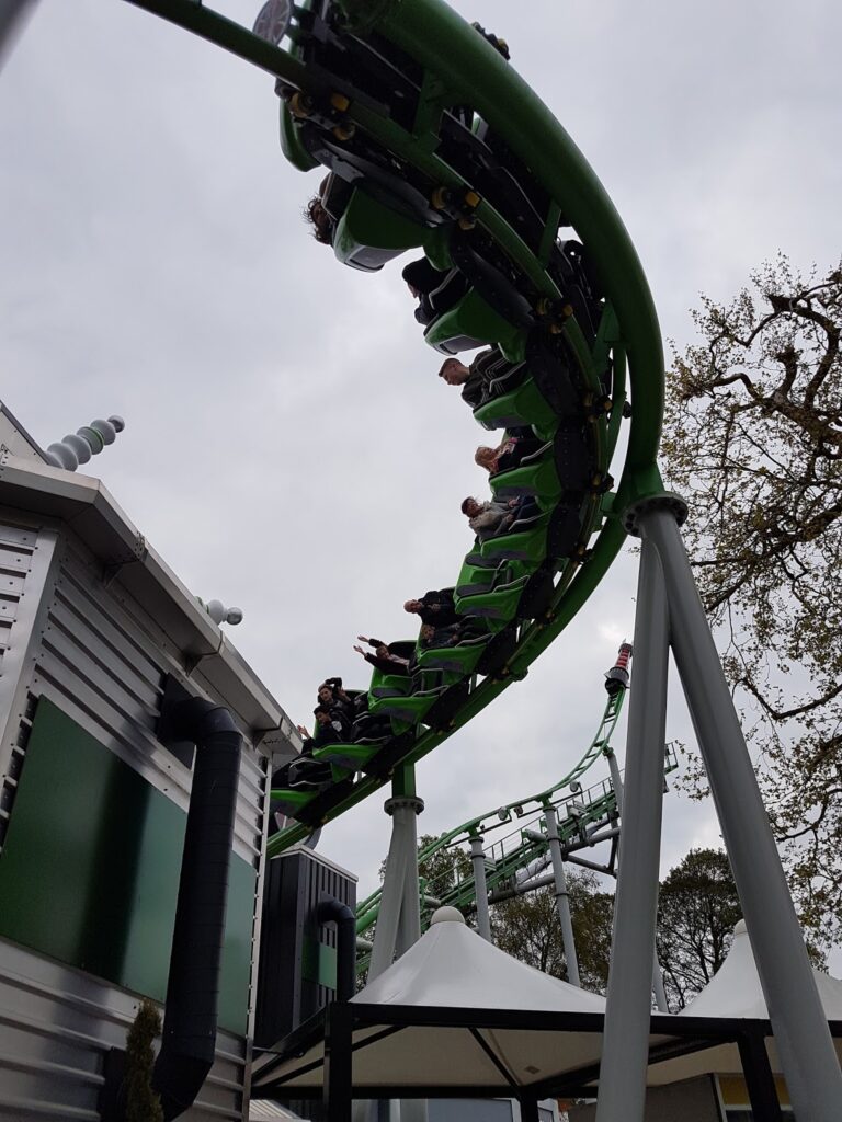 A green rollercoaster curves downward beside a metallic building. Riders with hands in the air appear excited. The sky is overcast, and trees with autumn leaves are visible in the background. A white canopy stands below the track.