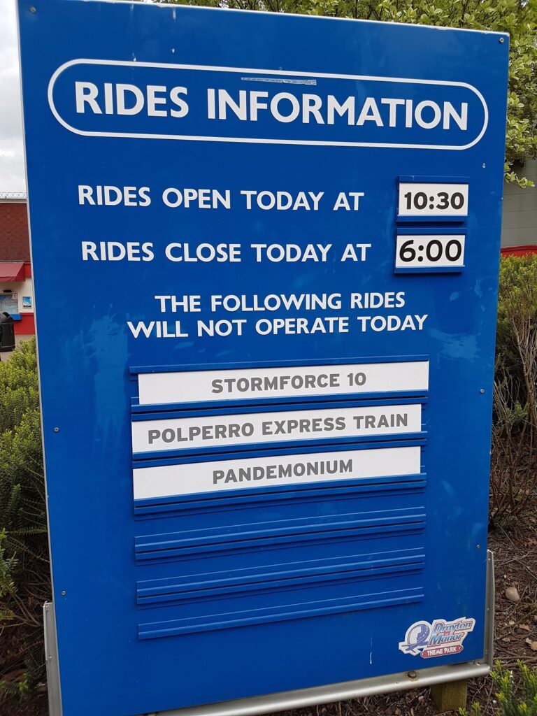 A blue sign reads "RIDES INFORMATION." Rides open at 10:30 and close at 6:00. Below, a notice lists rides not operating today: Stormforce 10, Polperro Express Train, and Pandemonium. Bushes and part of a red-roofed building are visible behind.