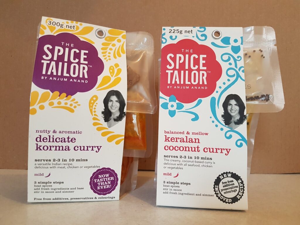 Two Spice Tailor curry kits stand side by side against a plain background. The left pack is "delicate korma curry" with a yellow accent, and the right is "keralan coconut curry" with a purple accent. Both feature a woman's photo and cooking details on the front.