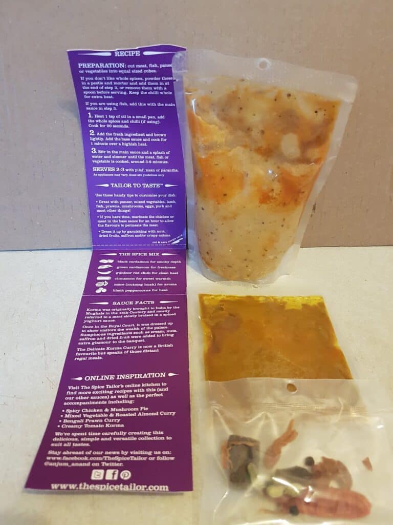 A packet of curry sauce, a small sachet of spices, and an opened instruction leaflet are displayed on a white surface. The leaflet is purple with white text, giving recipe directions and product information. The curry sauce is in a clear plastic pouch.