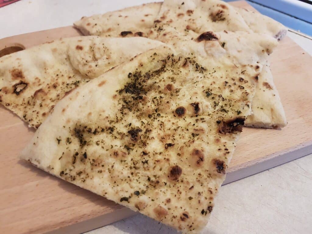 Several pieces of naan bread, lightly browned with charred spots and sprinkled with herbs, are arranged on a wooden chopping board. The bread appears soft and fluffy, with visible seasoning on the surface, placed on a light worktop.