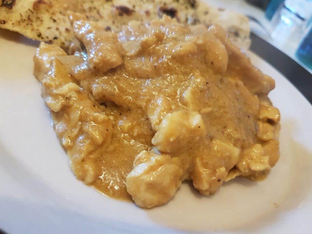 A close-up of creamy chicken curry with chunks of chicken in a thick golden-brown sauce, served on a white plate. Part of a piece of flatbread is visible in the background. The dish looks rich and flavourful.