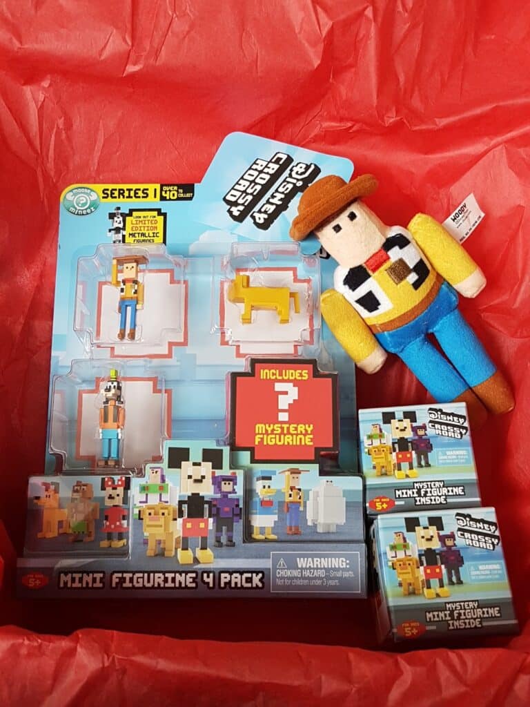 A gift box with red tissue paper contains a Disney Pixar Toy Story Woody soft toy, a blue-packaged 4-pack of Minecraft-style Disney mini figures, and two small boxes labelled “Disney Mystery Figure.” The figures and packaging are colourful and pixelated.