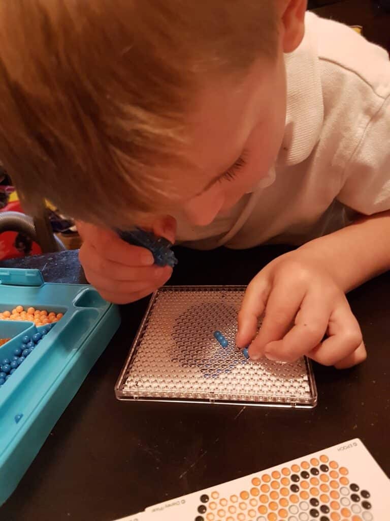 A young child in a white shirt leans closely over a clear pegboard, carefully placing blue beads onto it with one hand, whilst holding more beads in the other. A tray of assorted beads and a pattern card are nearby on the table.