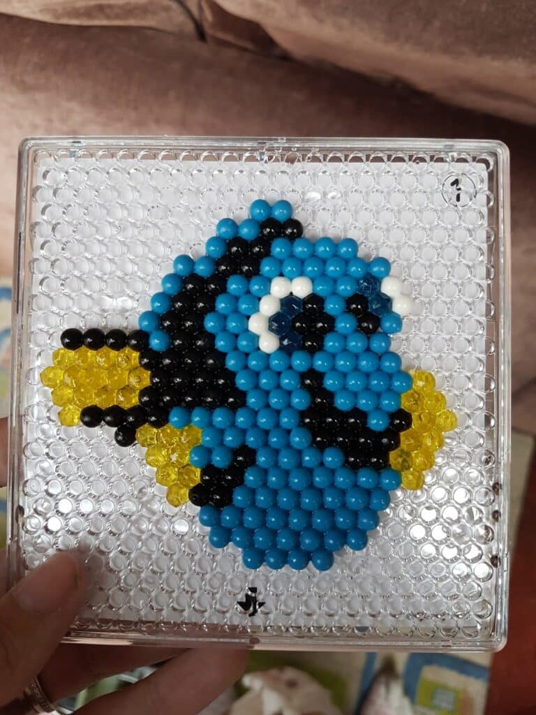 A hand holds a square pegboard with colourful beads arranged to depict Dory, the blue tang fish from “Finding Nemo,” featuring blue, black, white, and yellow beads to resemble her distinct features. The background is transparent with visible pegs.
