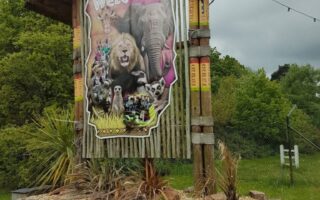 A large wooden sign reads "Welcome" and features images of wild animals, including a lion, giraffe, zebra, and monkeys. The sign stands outdoors by a road, surrounded by greenery, with part of a red car visible in the bottom right corner.