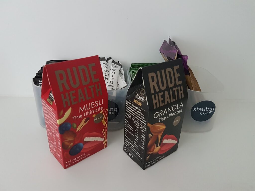 Two colourful cardboard boxes labelled "RUDE HEALTH"—one red for muesli, one black for granola—stand in front of a grey container filled with various single-serve coffee and tea packets. The container has a blue "staying cool" label. All items are on a white surface.