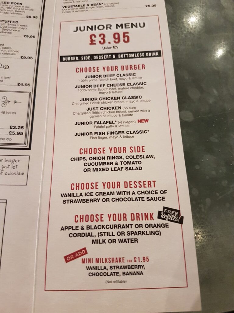 A restaurant junior menu offers a burger, side, dessert, and bottomless drink for £3.95. Options include beef, chicken, fish, or falafel, with sides, desserts, and drinks listed. A mini milkshake is available for £1.95. The menu is printed on a table top.