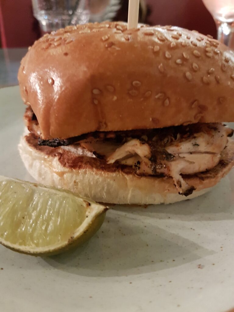 A grilled chicken sandwich with a sesame seed bun is served on a plate next to a wedge of lime. The sandwich is held together with a wooden skewer, and a glass is blurred in the background.