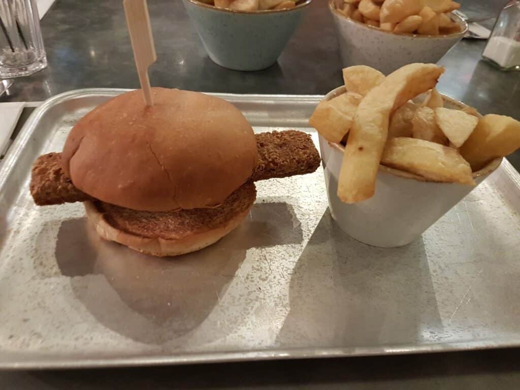 A sandwich with a breaded, rectangular fried item in a bun sits on a metal tray next to a cup of chunky chips. Two more bowls of chips and glasses of water are visible in the background on the table.