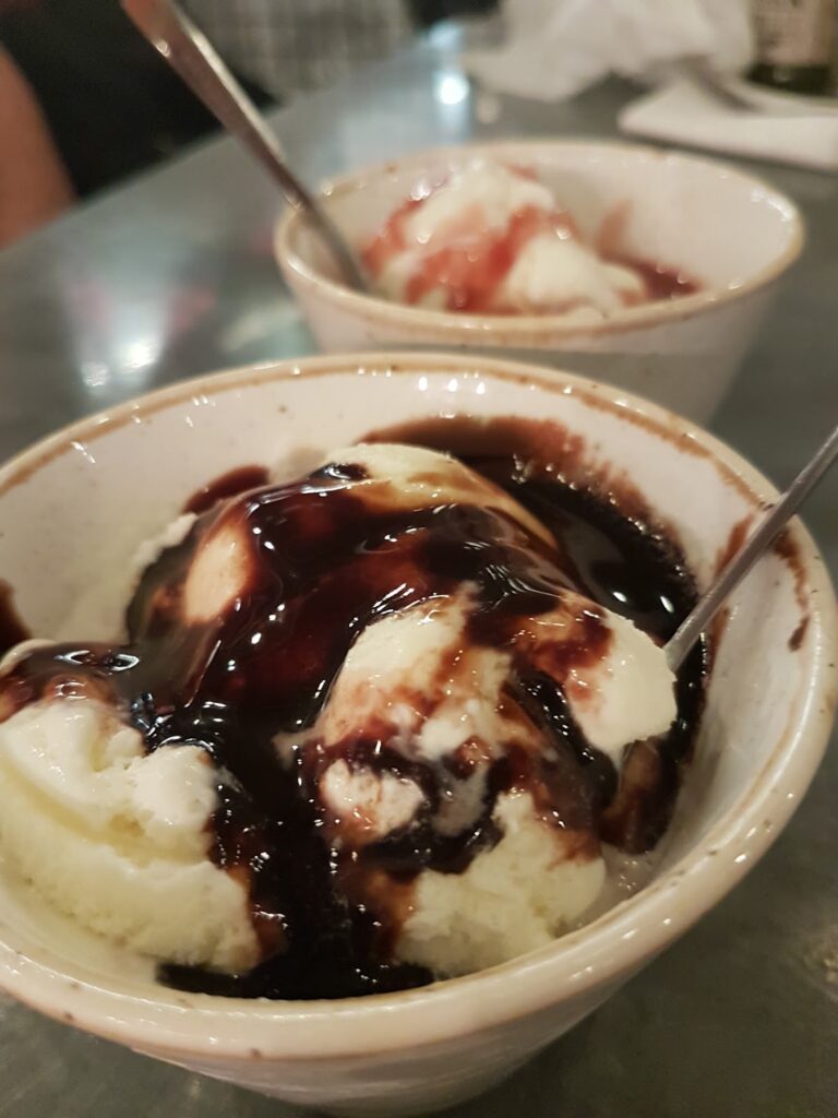 Two white bowls of vanilla ice cream are topped with chocolate sauce in the front bowl and strawberry sauce in the bowl behind it. Each bowl has a metal spoon, and both are placed on a reflective surface with a blurred background.
